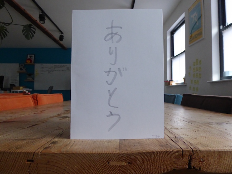 A white card with the word ‘thank you’ hand-written in Japanese hiragana characters stood on a wooden table.